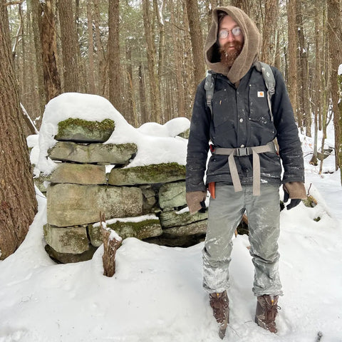 A bearded wizard wearing a dark hooded jacket, gray pants, and brown hiking boots stands in a snowy forest next to a mossy stone wall preparing to harvest some chaga for his potions.